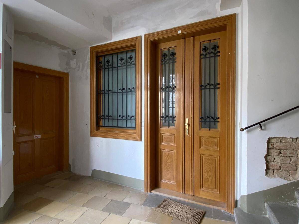 Eingangsbereich mit Holztüren und einem Fenster mit Gitter, gefliester Boden und Treppenaufgang.