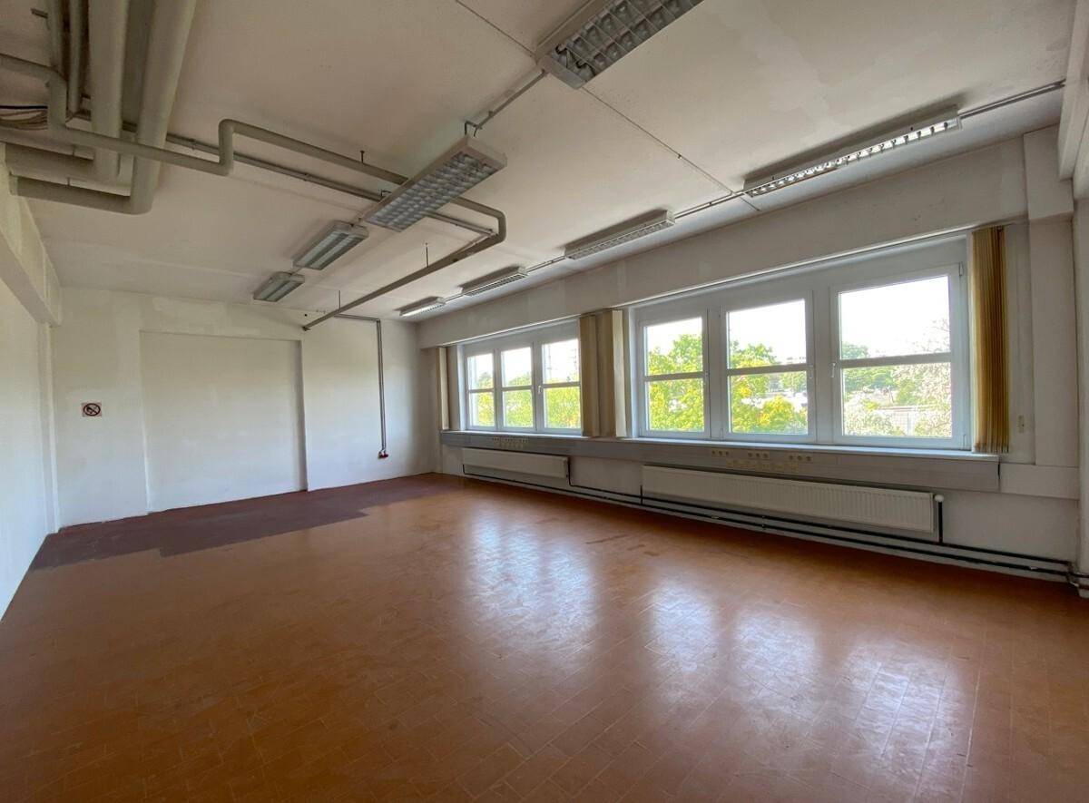 Geräumiger Büroraum mit großen Fenstern, sichtbaren Leitungen an der Decke und Fliesenboden.