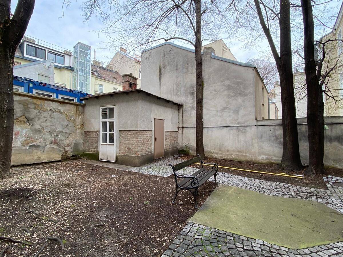 Ruhiger Hinterhof oder Gartenbereich mit kahlen Bäumen, einer Bank und einem kleinen Nebengebäude.