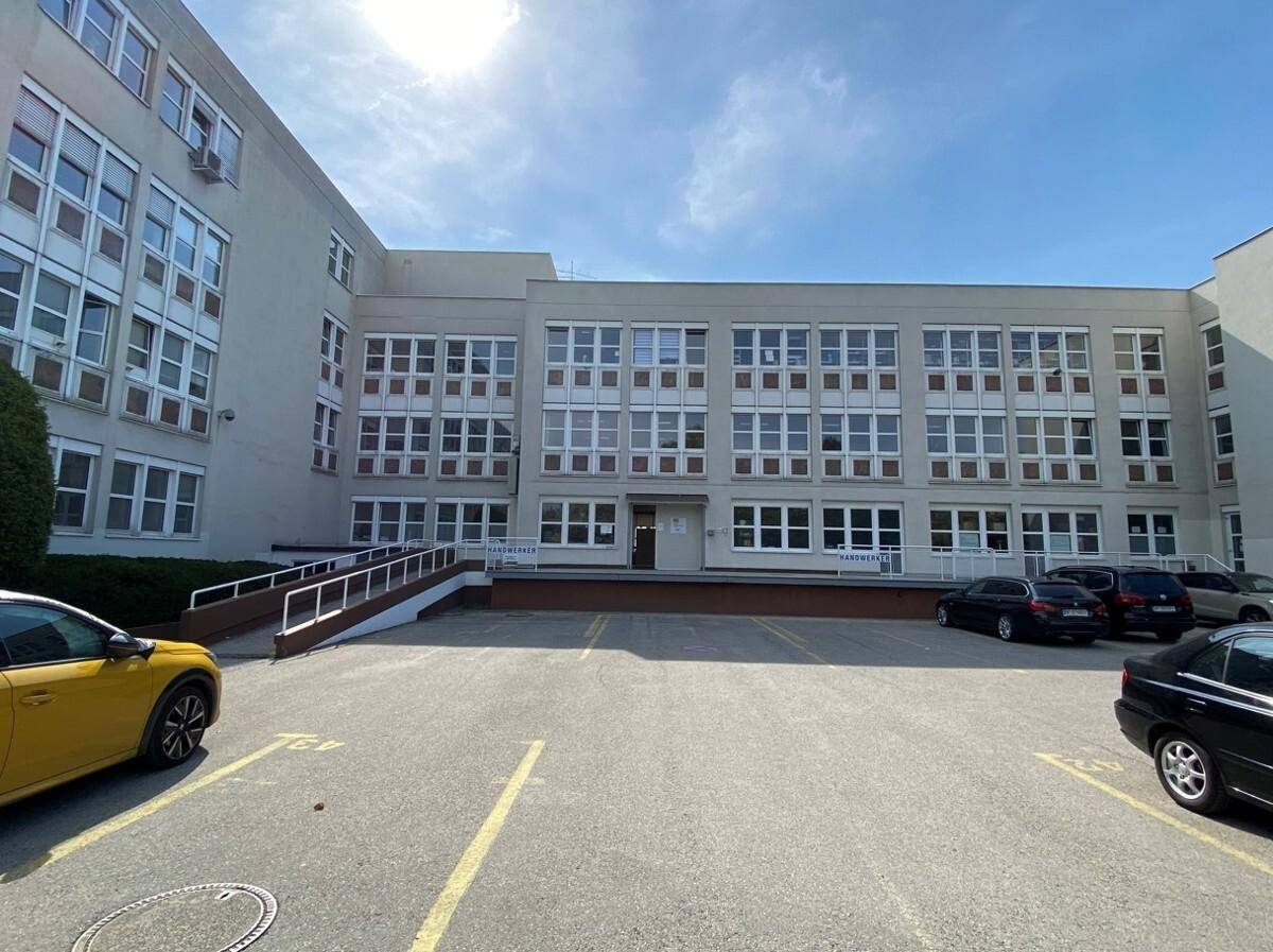 Vorderansicht eines großen Gebäudes mit weitläufigem Parkplatz und einer barrierefreien Rampe zum Eingang.