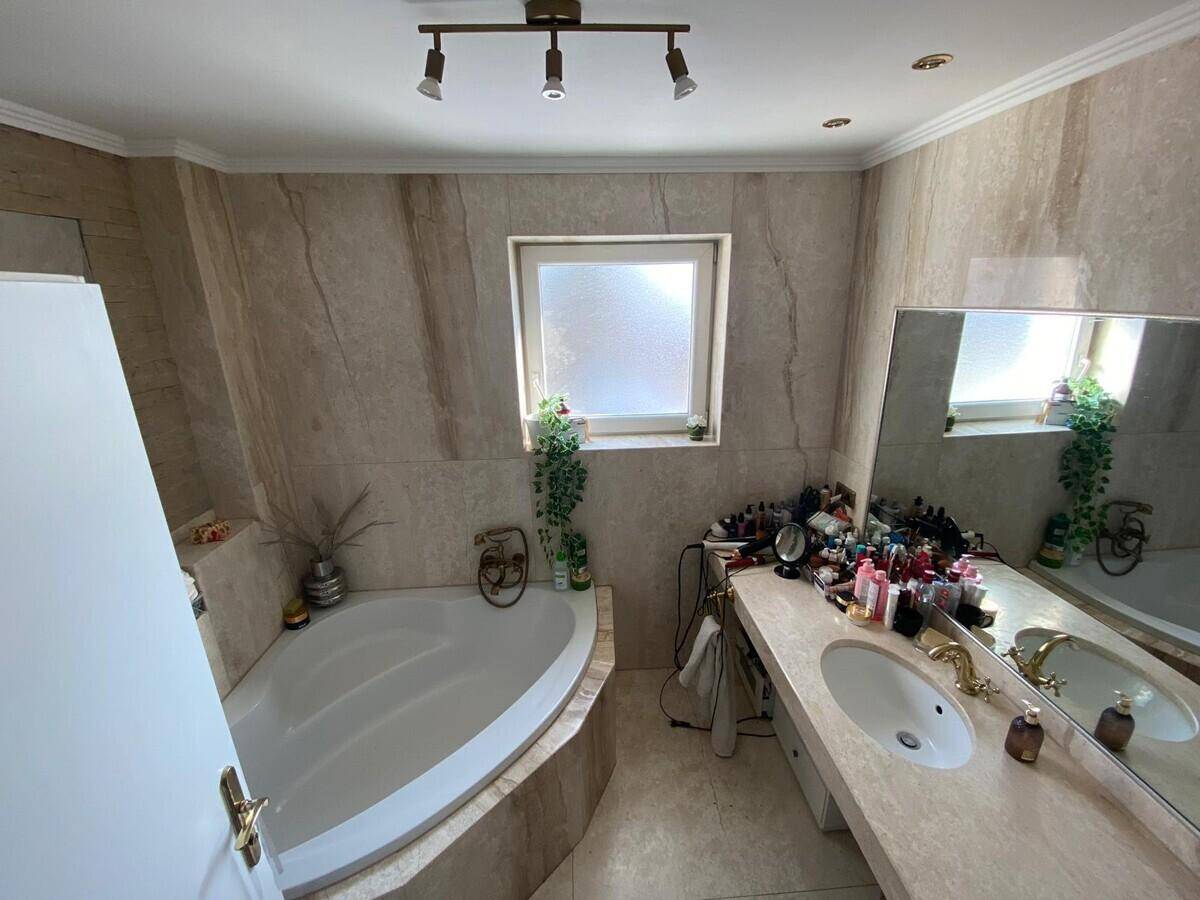 Helles Badezimmer mit Eckbadewanne, großem Waschtisch und Fenster, alles in beigen Marmorfliesen gehalten.