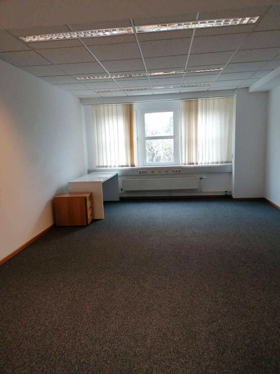Leeres Büro mit grauem Teppichboden, einem Fenster mit Jalousien und einem kleinen Schreibtisch.