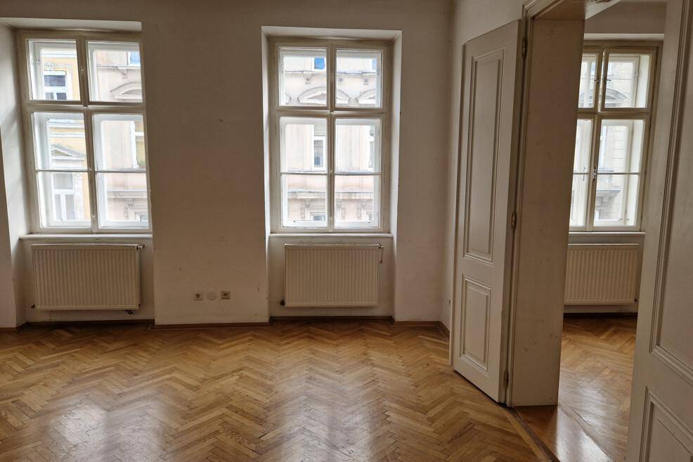 Geräumiges Zimmer mit Fischgrätparkett, hohen Fenstern und einer Doppeltür, typisch für einen Altbau.