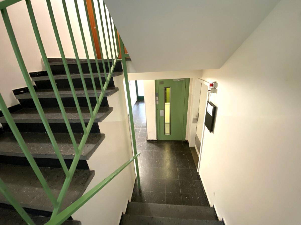 Treppenhaus mit dunklen Stufen und grünem Geländer, ein Aufzug ist im Hintergrund sichtbar.