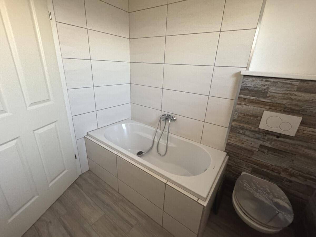 Modernes Badezimmer mit weiß gefliester Badewanne und wandhängendem WC, akzentuiert durch Holzoptikfliesen.