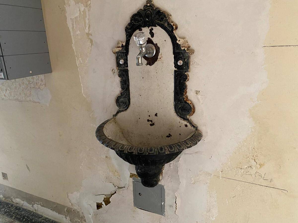 Historisches, verziertes Wandwaschbecken in einem Flur mit sichtbaren Abnutzungsspuren und beschädigter Wand.