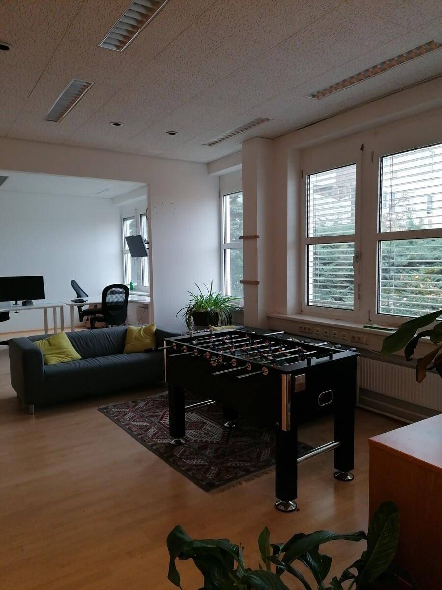 Heller Büroraum mit Holzboden, großen Fenstern und einem Kicker-Tisch, der eine entspannte Arbeitsatmosphäre bietet.