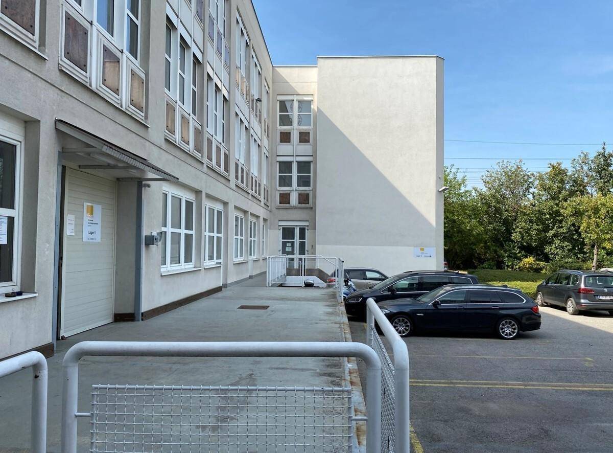 Außenansicht eines modernen Gebäudes mit Parkplätzen und grüner Umgebung unter blauem Himmel.