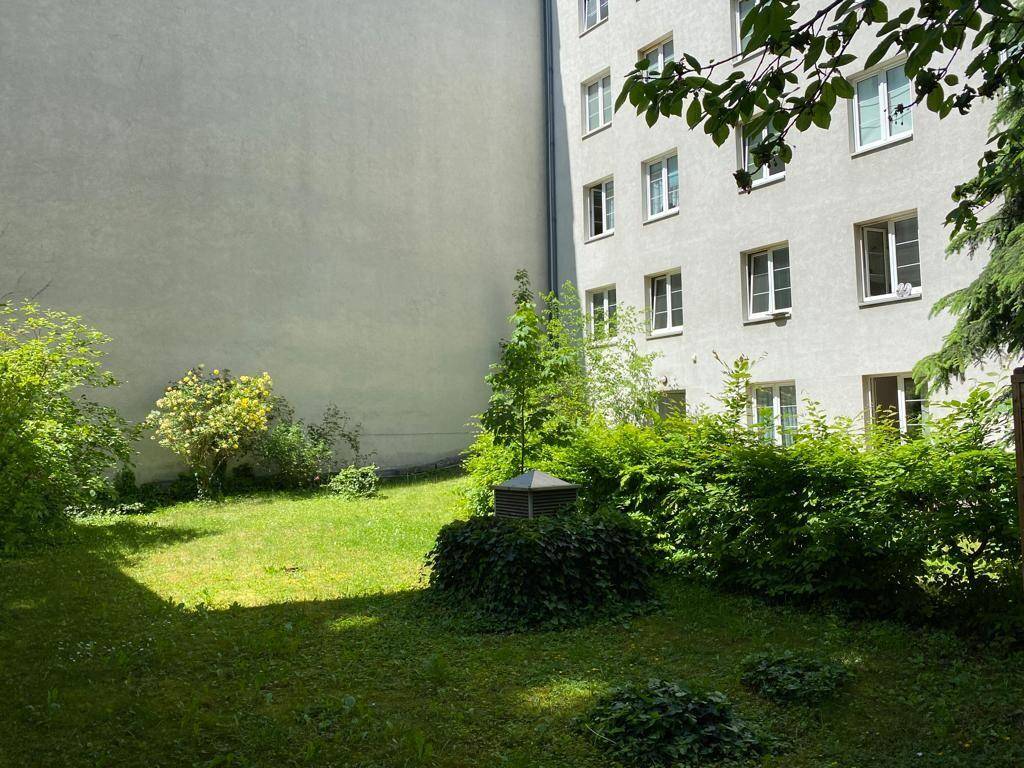 Grüner Innenhofgarten mit Rasenfläche und Sträuchern, umgeben von Gebäudewänden.