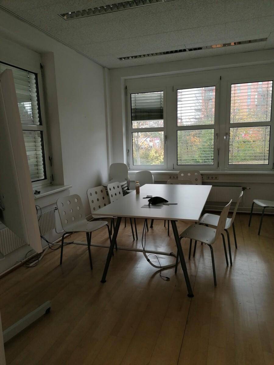 Heller Büroraum mit Holzboden, einem Konferenztisch und großen Fenstern mit Jalousien.