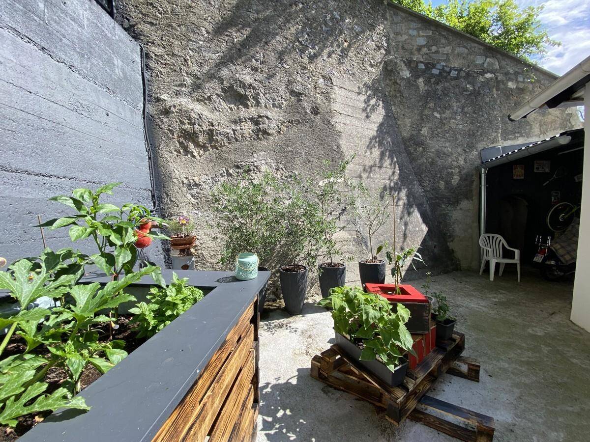 Kleiner, gepflasterter Gartenbereich mit Hochbeeten und Topfpflanzen vor einer hohen Steinmauer.