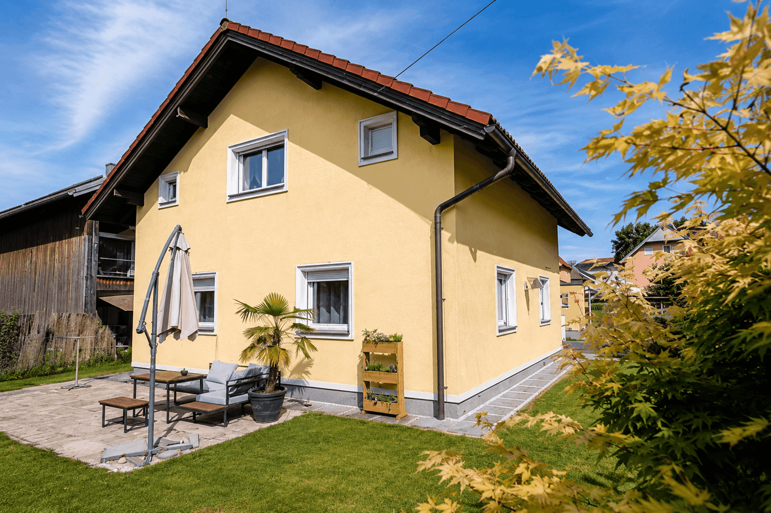 Seitenansicht des Hauses mit gepflasterter Terrasse, Gartenmöbeln und kleiner Rasenfläche.