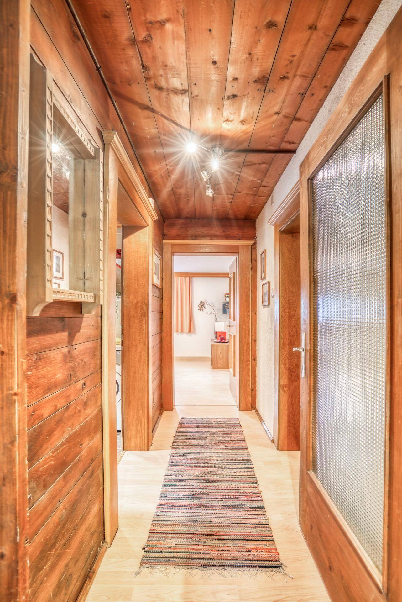 Rustikaler Flur mit Holzwänden und Holzdecke, beleuchtet von Deckenspots und einem bunten Teppich.