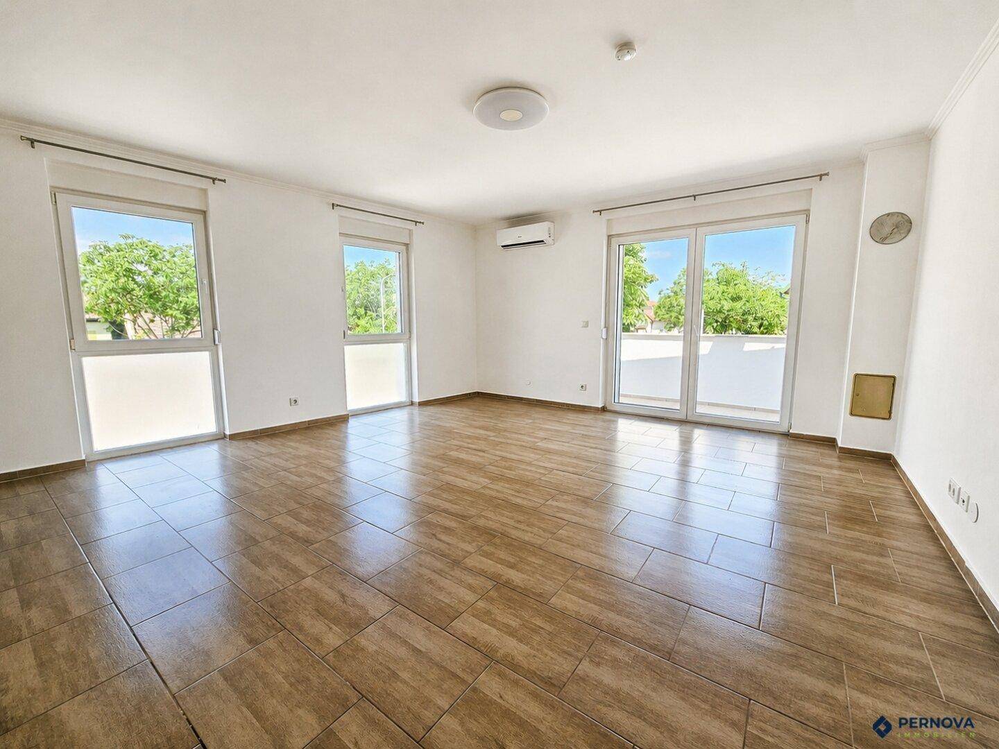 Helles, leeres Wohnzimmer mit großen Fenstern, Fliesenboden und Zugang zum Balkon.