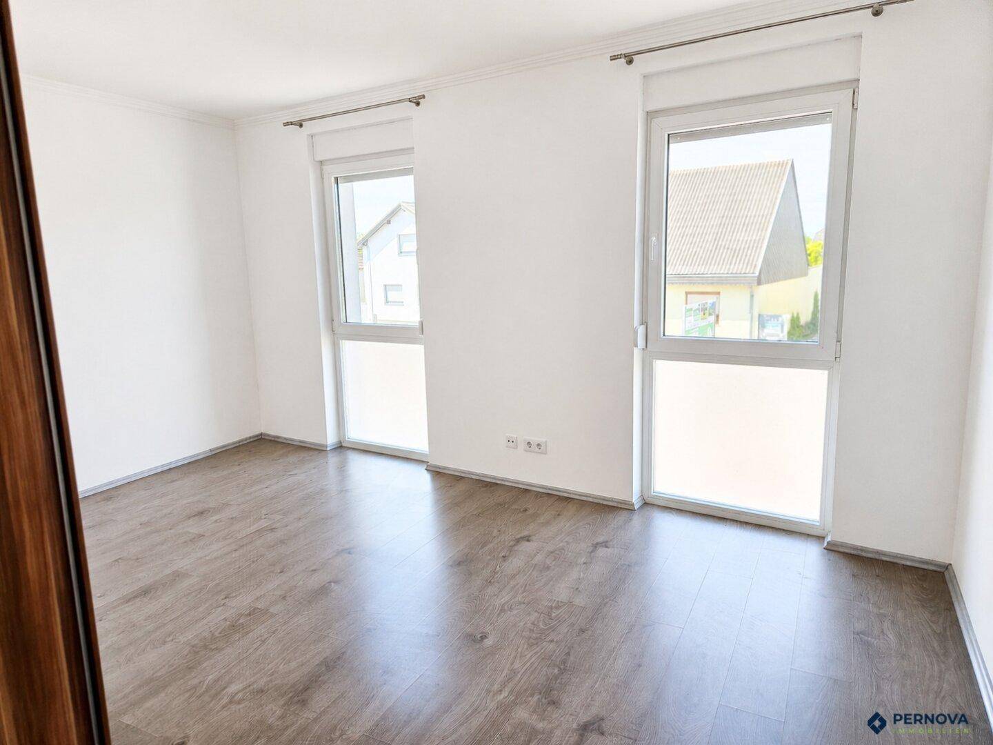Geräumiges Schlafzimmer mit zwei großen Fenstern und dunklem Laminatboden.