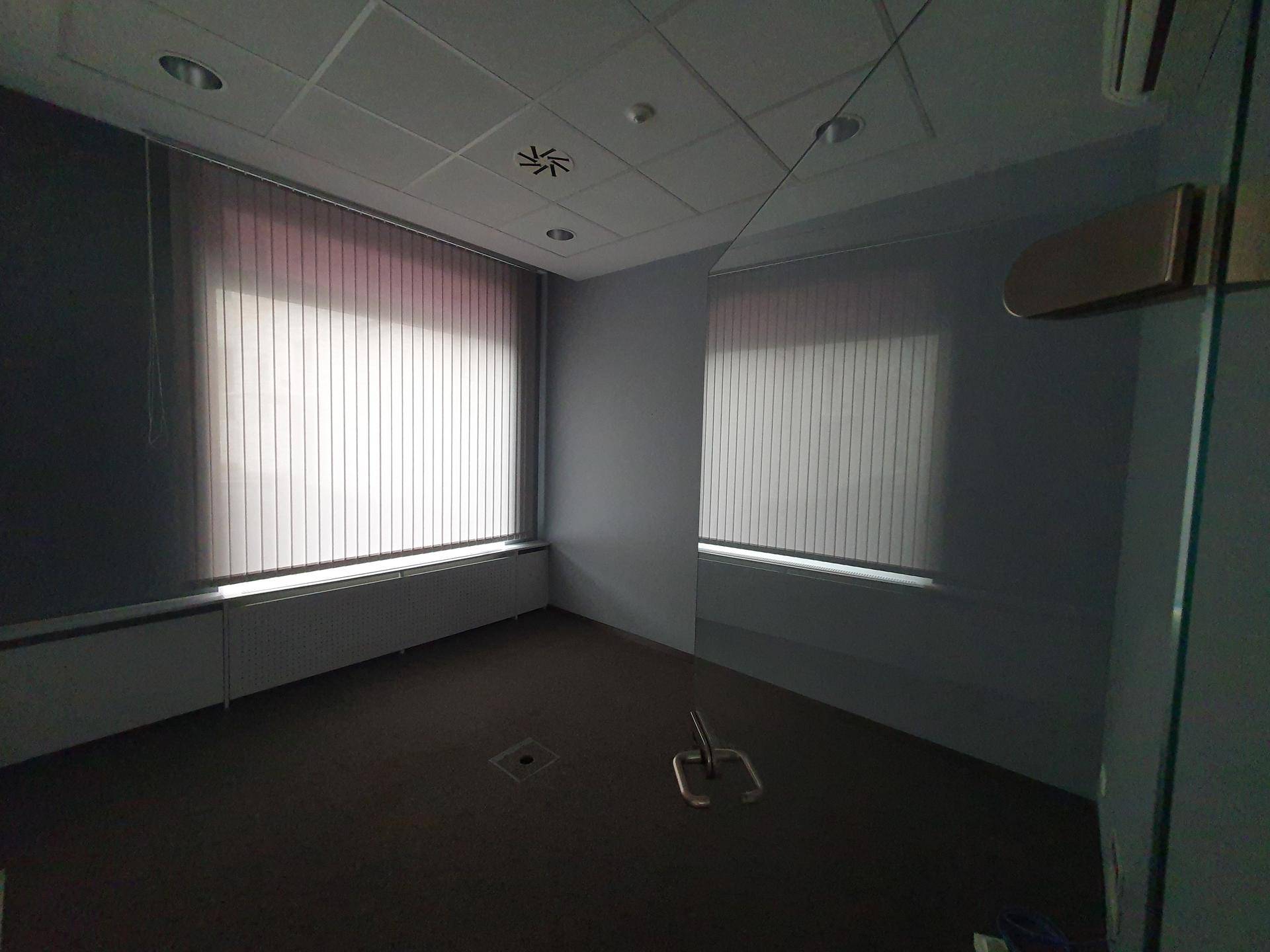 Kleiner Büroraum mit Teppichboden und vertikalen Jalousien an den Fenstern.