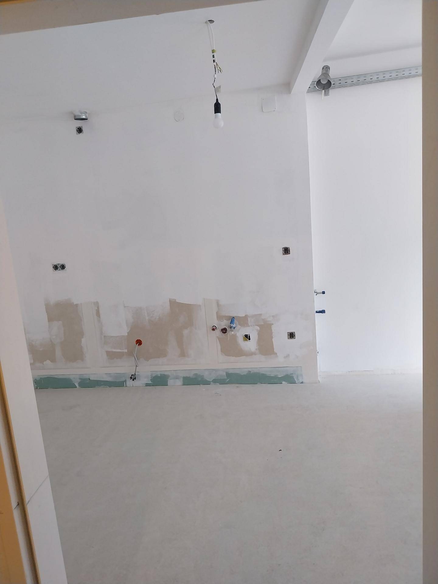 Raum im Bau mit vorbereiteten Anschlüssen für eine Küche und provisorischer Beleuchtung.