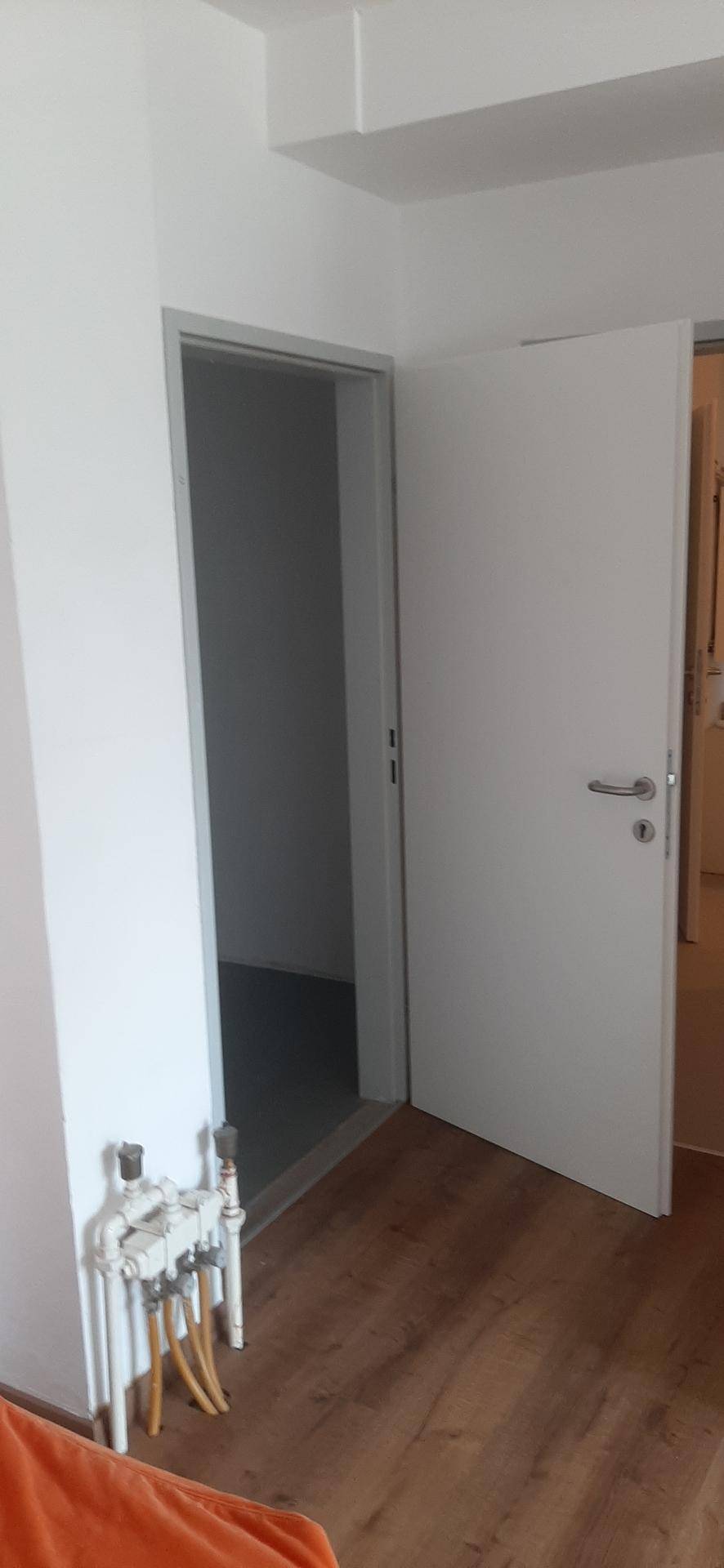 Flur mit offener Tür und sichtbaren Rohrleitungen an der Wand, auf dunklem Laminatboden.