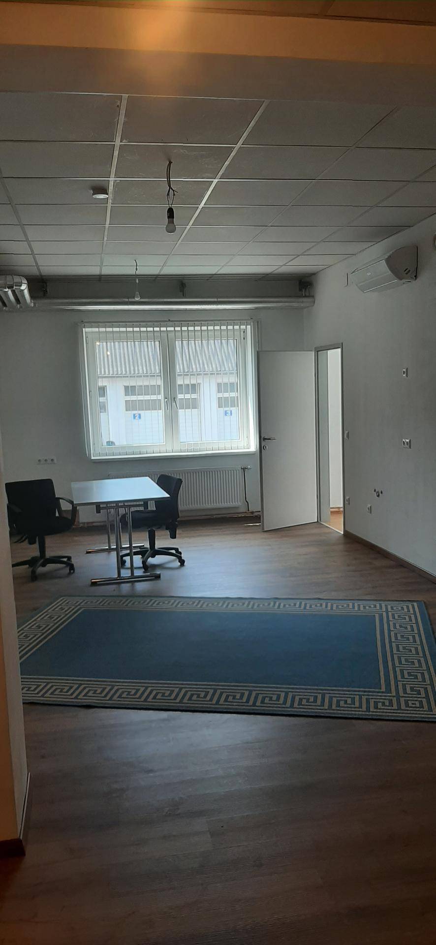 Geräumiger Büroraum mit dunklem Laminatboden, Fensterfront, Klimaanlage und einem blauen Teppich.