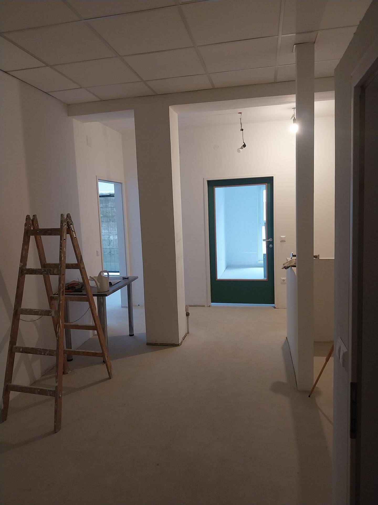 Offener Raum im Bau mit Säulen, einer Leiter und einer grünen Eingangstür.