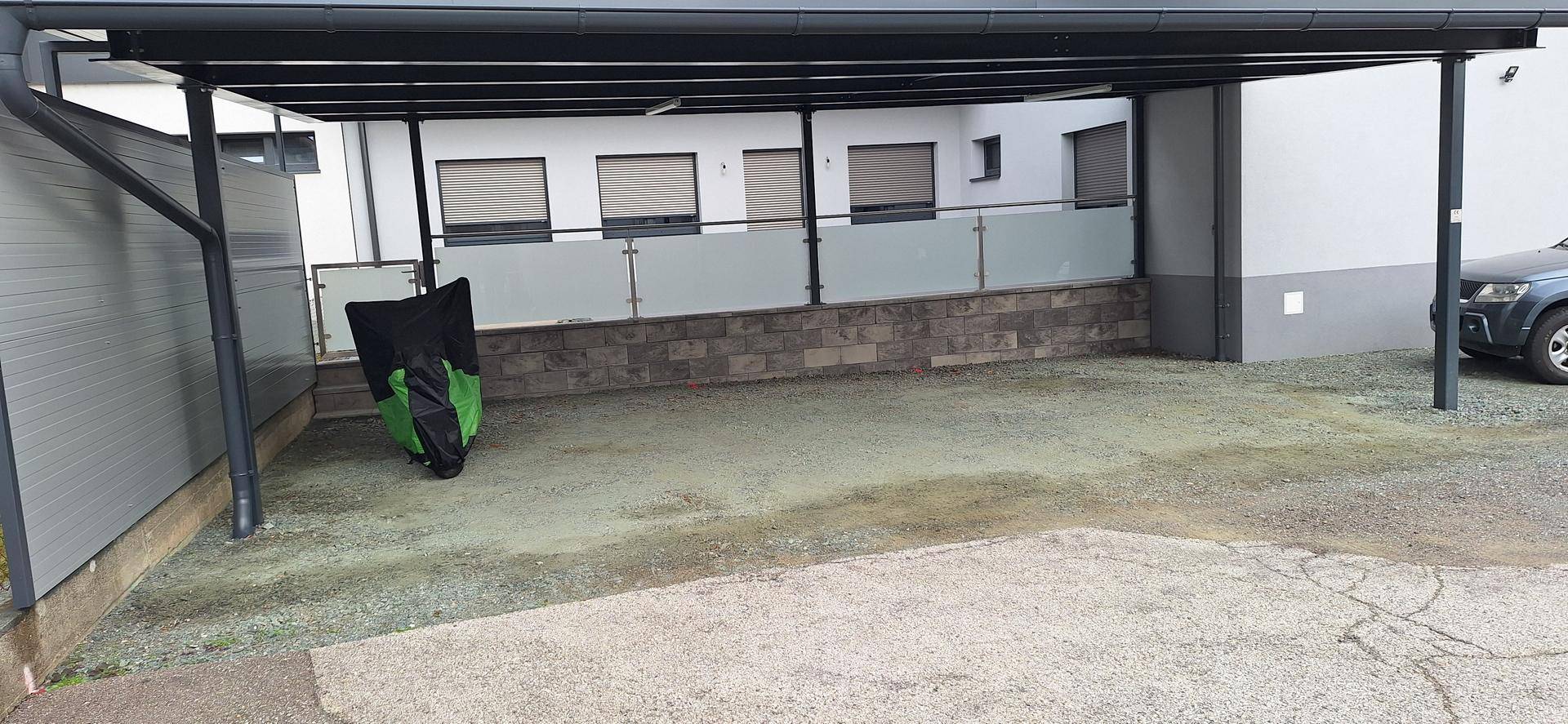 Überdachter Stellplatz (Carport) mit Kiesfläche und Blick auf die moderne Gebäudefassade im Hintergrund.