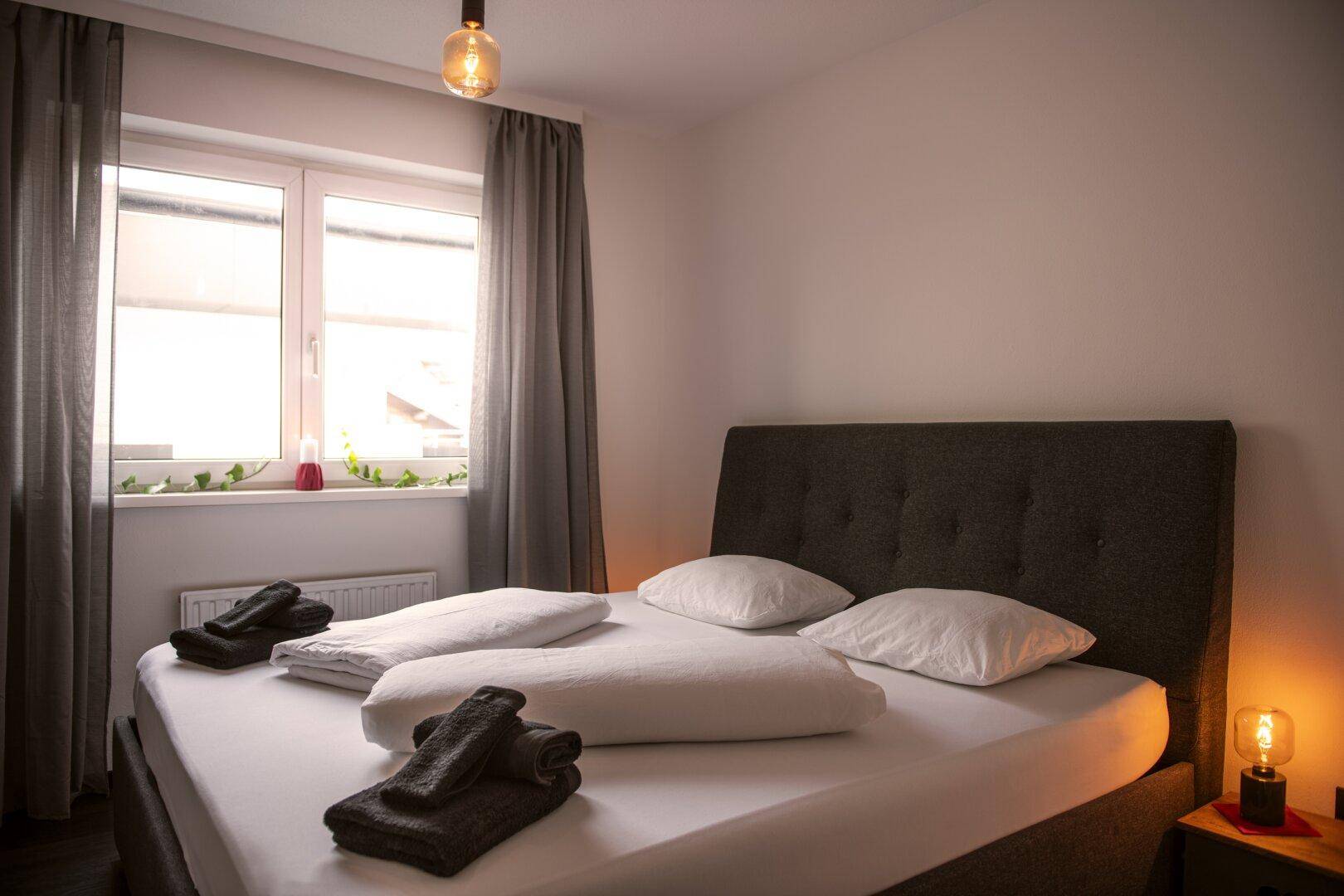 Helles Schlafzimmer mit Doppelbett, Fenster und schlichten Vorhängen, gemütlich beleuchtet.