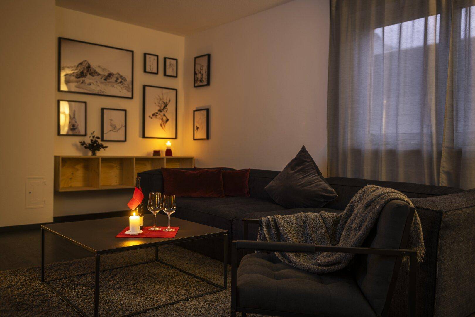 Gemütliches Wohnzimmer mit dunklem Sofa, Sessel und stimmungsvoller Beleuchtung, dekoriert mit Bergmotiven.