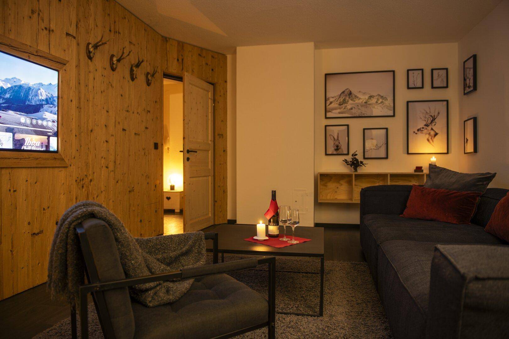 Gemütliches Wohnzimmer mit dunklem Sofa, Holzwand und stimmungsvoller Beleuchtung, mit Bergbildern.