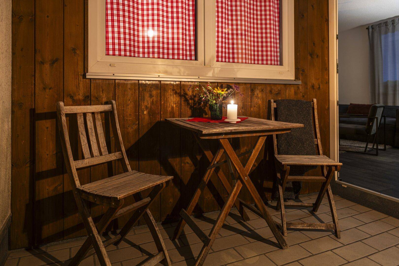 Kleiner Balkon mit Holztisch und Stühlen, beleuchtet von einer Kerze, mit karierten Vorhängen.