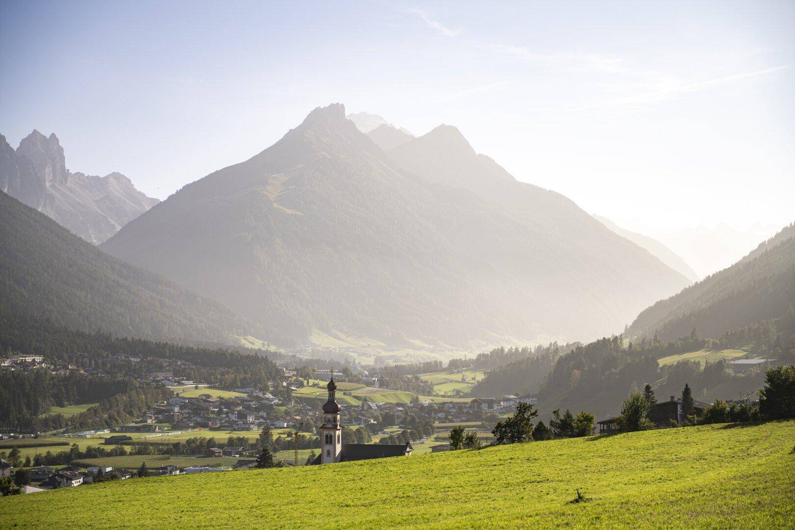 Malerische Berglandschaft mit einem Dorf und einer Kirche im Tal, umgeben von grünen Wiesen.