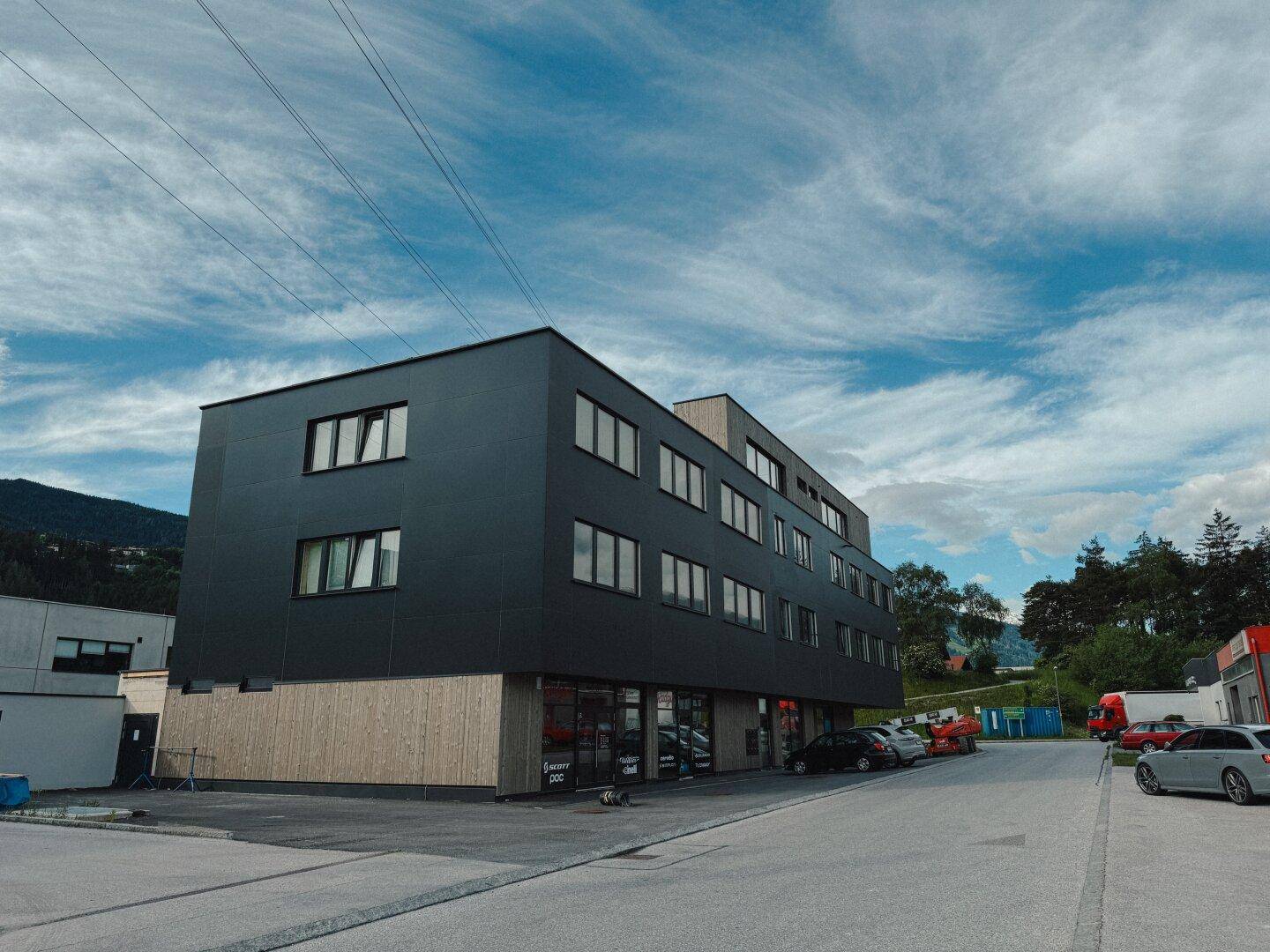 Modernes Gebäude mit dunkler Fassade und Holzverkleidung im Erdgeschoss, umgeben von Parkplätzen und Bergen.