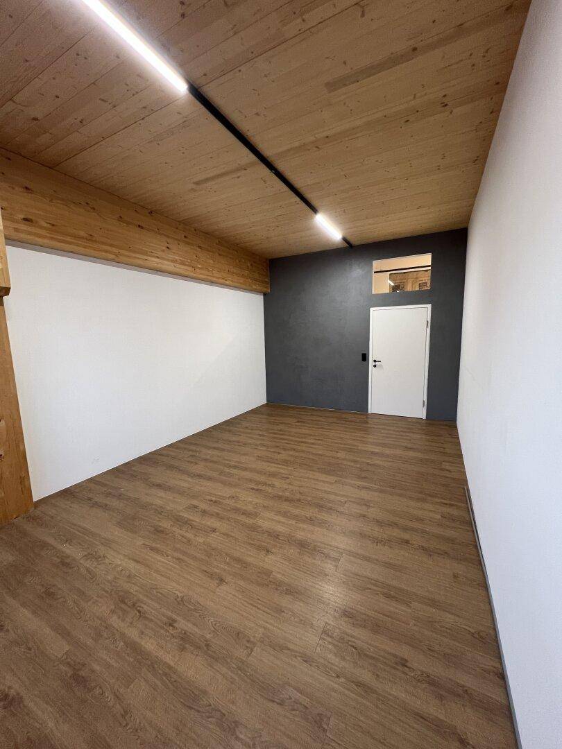 Moderner Raum mit Holzbalkendecke, weißen Wänden und einer Akzentwand in Dunkelgrau, ausgestattet mit Laminatboden.