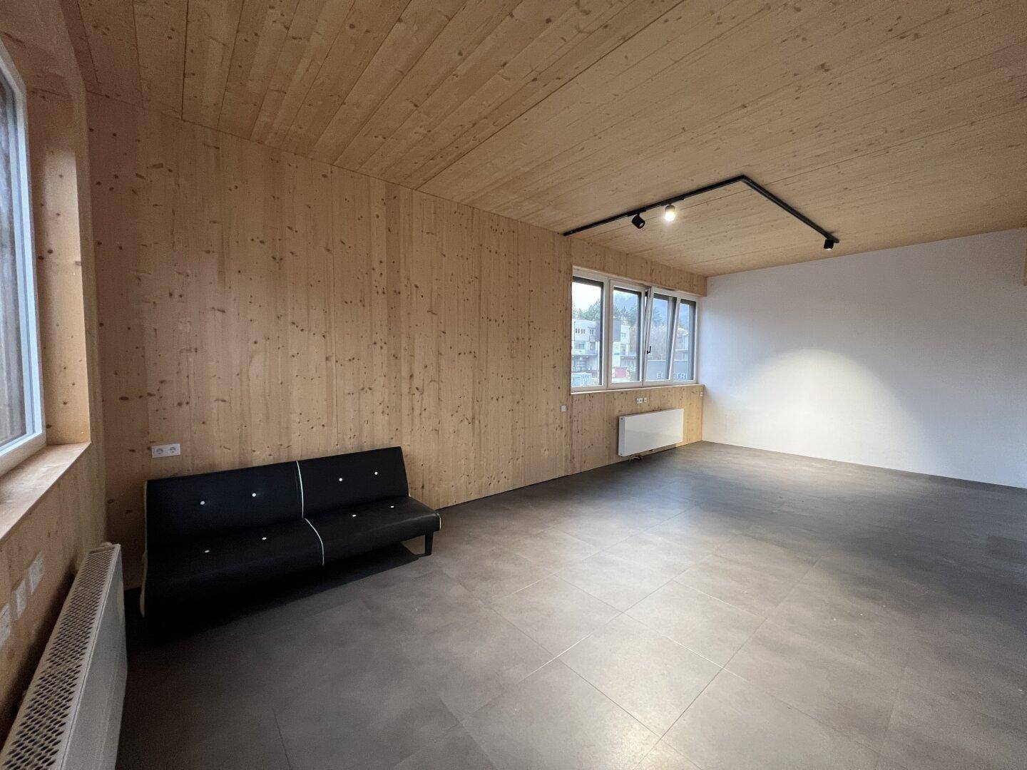 Moderner Raum mit dunklem Fliesenboden, Holzwänden und -decke, ausgestattet mit einem schwarzen Sofa und Fenstern.