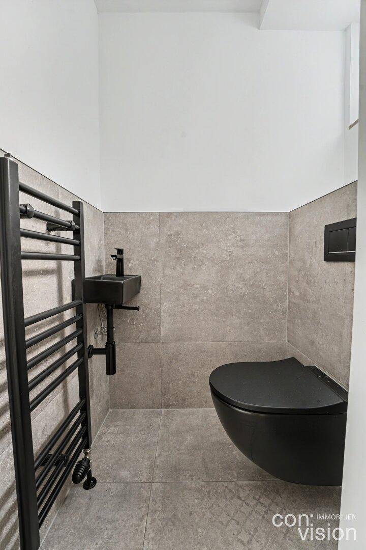 Modernes Gäste-WC mit schwarzer Toilette, kleinem Waschbecken und beheiztem Handtuchhalter.