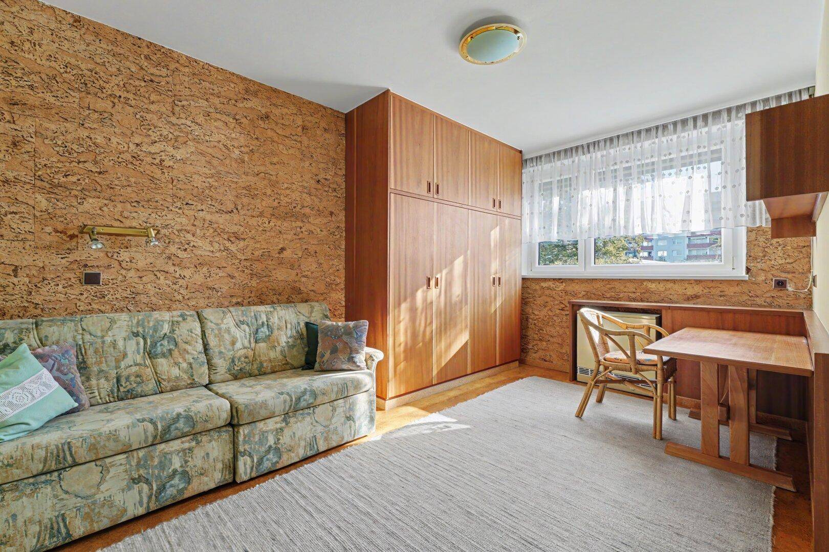 Schlafzimmer mit Korktapete, Holzschrank, Schlafsofa und kleinem Arbeitsbereich am Fenster.