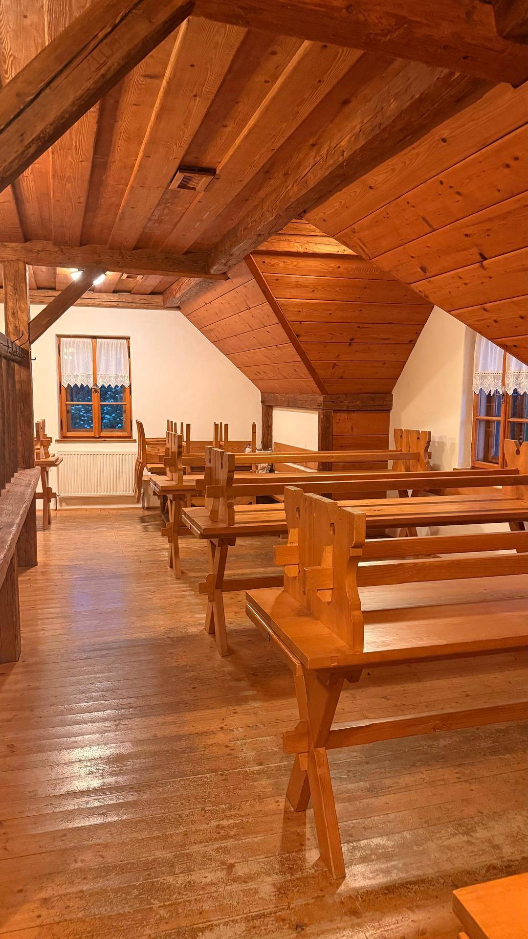 Großer Dachgeschossraum mit schräger Holzdecke und Reihen von rustikalen Holzbänken und Tischen.