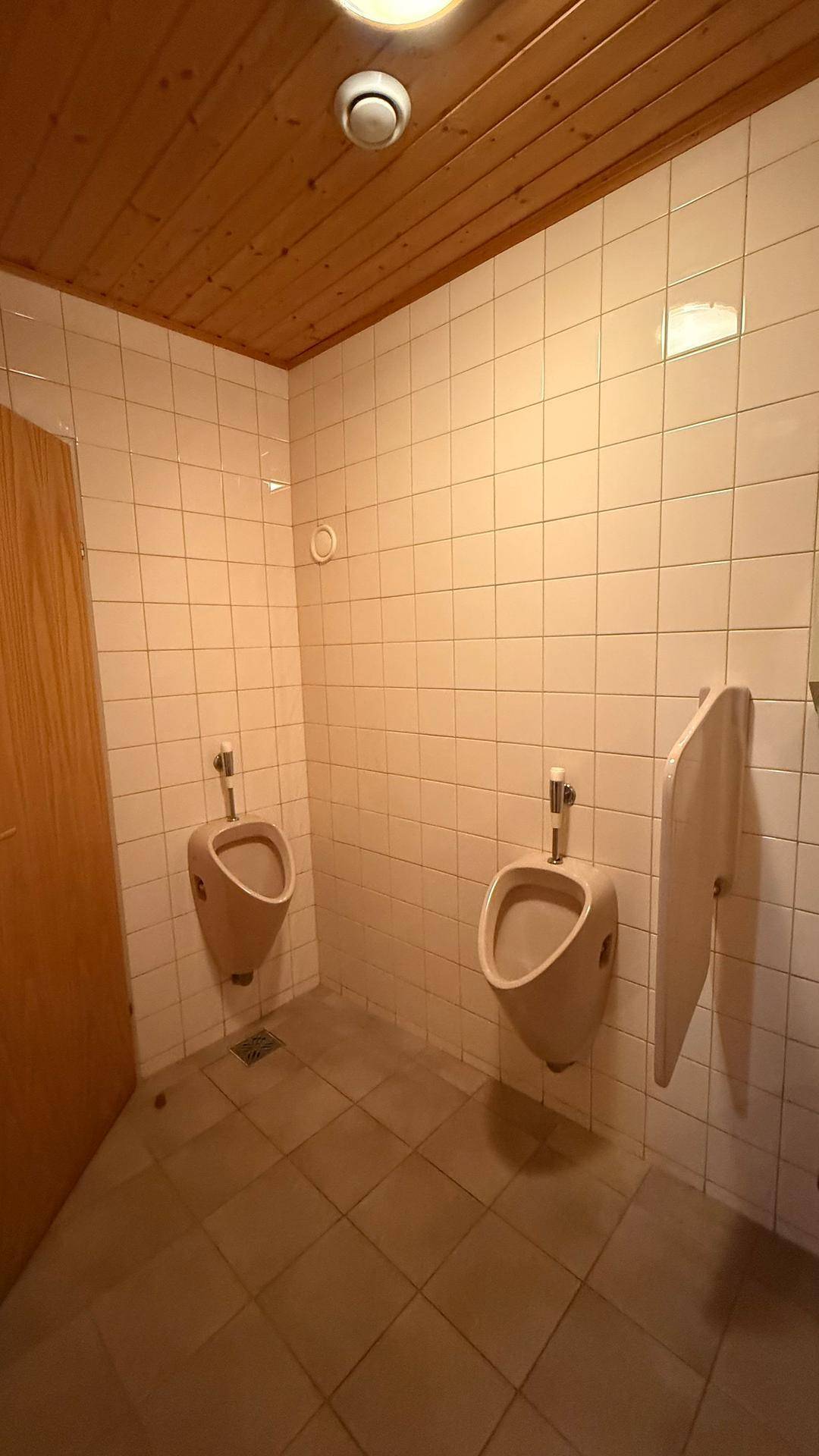 Herren-WC mit zwei Urinalen, hell gefliesten Wänden und einem Decke aus Holzpaneelen.