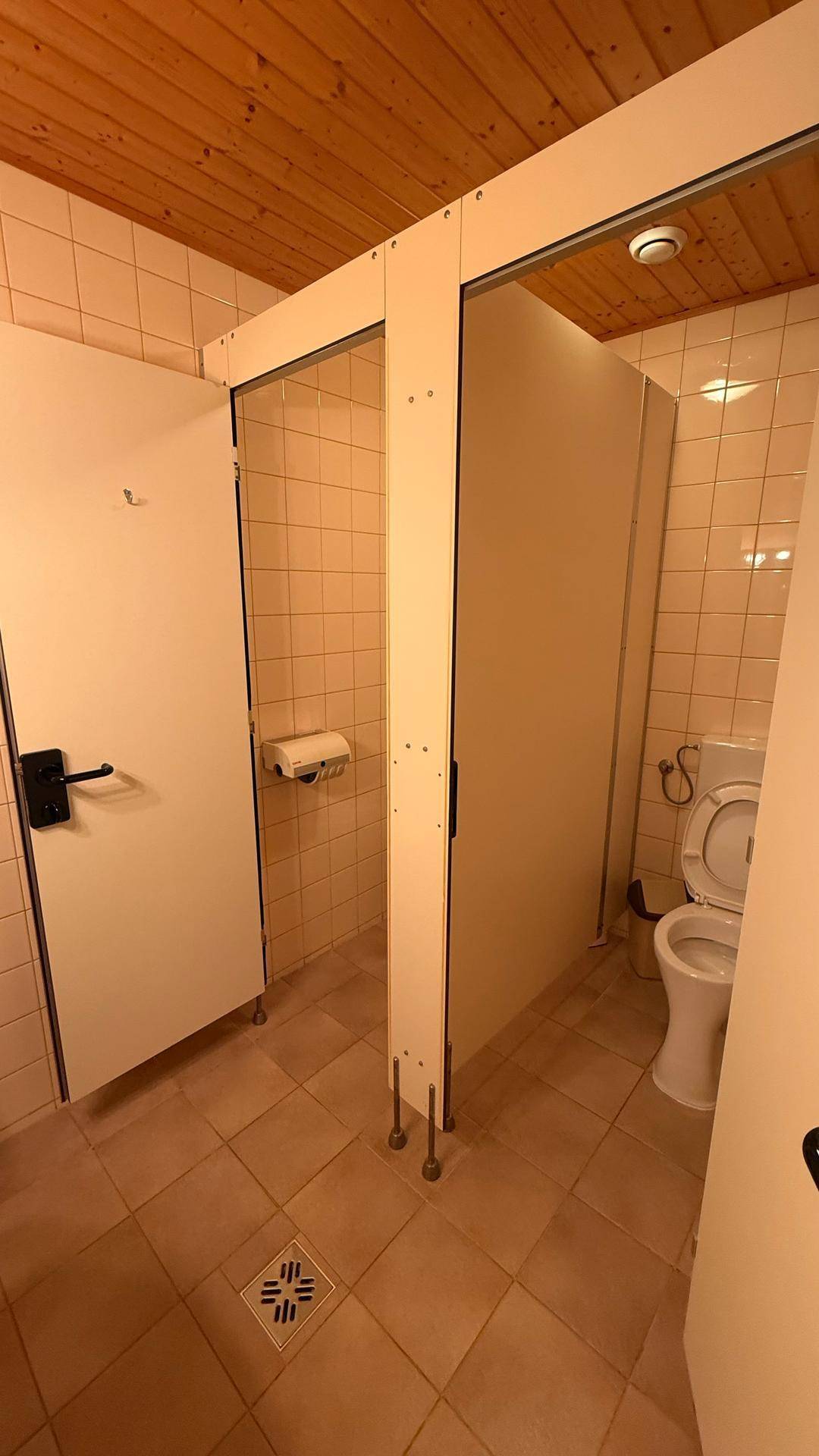 Gefliester Toilettenbereich mit mehreren Kabinen, einem WC und einer Holzpaneeldecke.