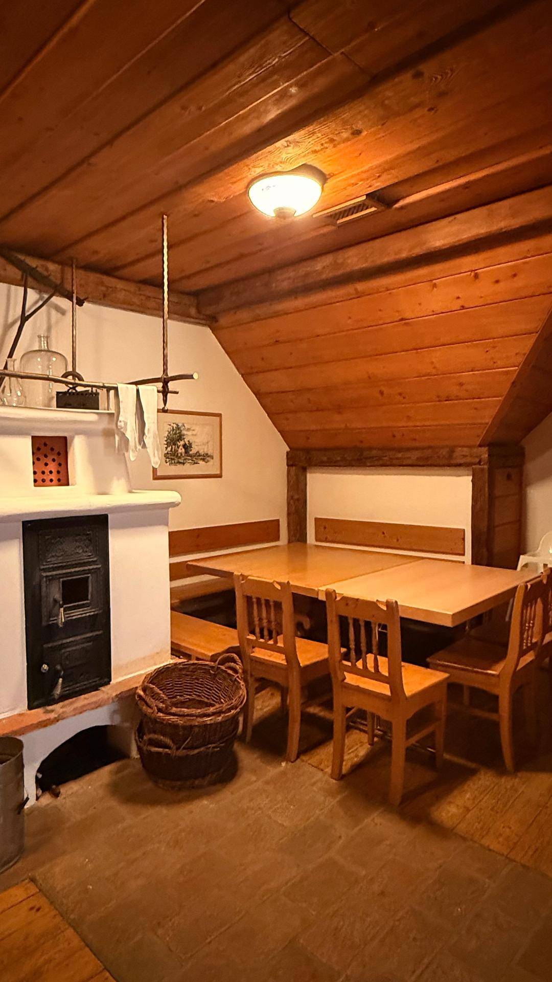 Dachgeschoss-Essbereich mit schräger Holzdecke, einem traditionellen Kachelofen und Holztischen.