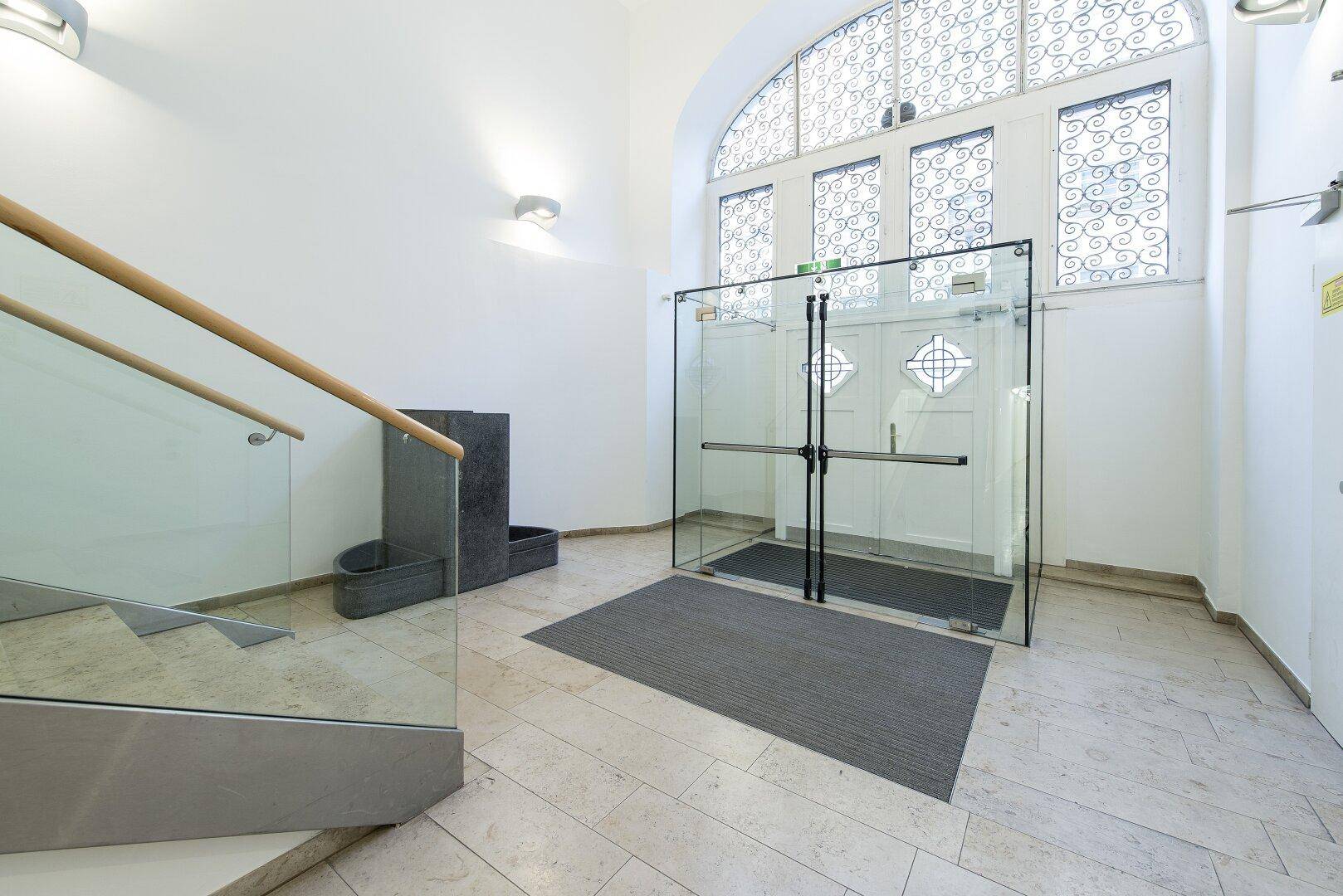 Heller Eingangsbereich mit hohen Decken, moderner Glastreppe und großen Fenstern. Der Boden ist mit hellen Steinfliesen ausgelegt.