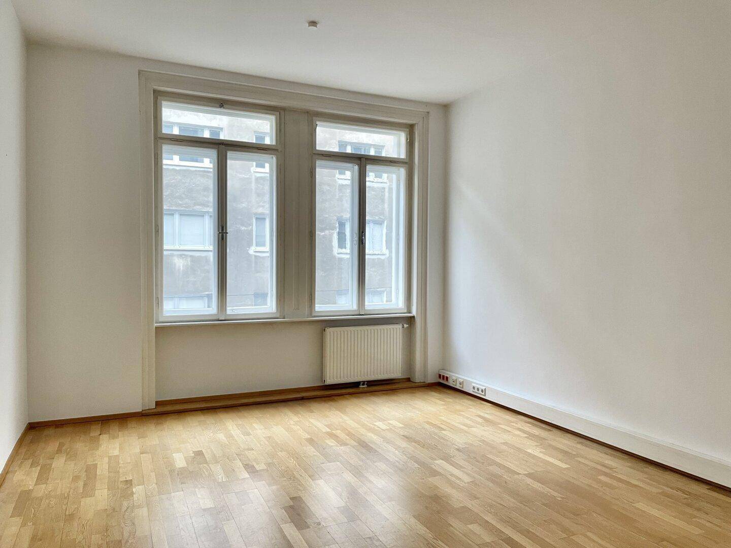 Heller Raum mit zwei großen Fenstern, Parkettboden und Heizkörper unter den Fenstern.