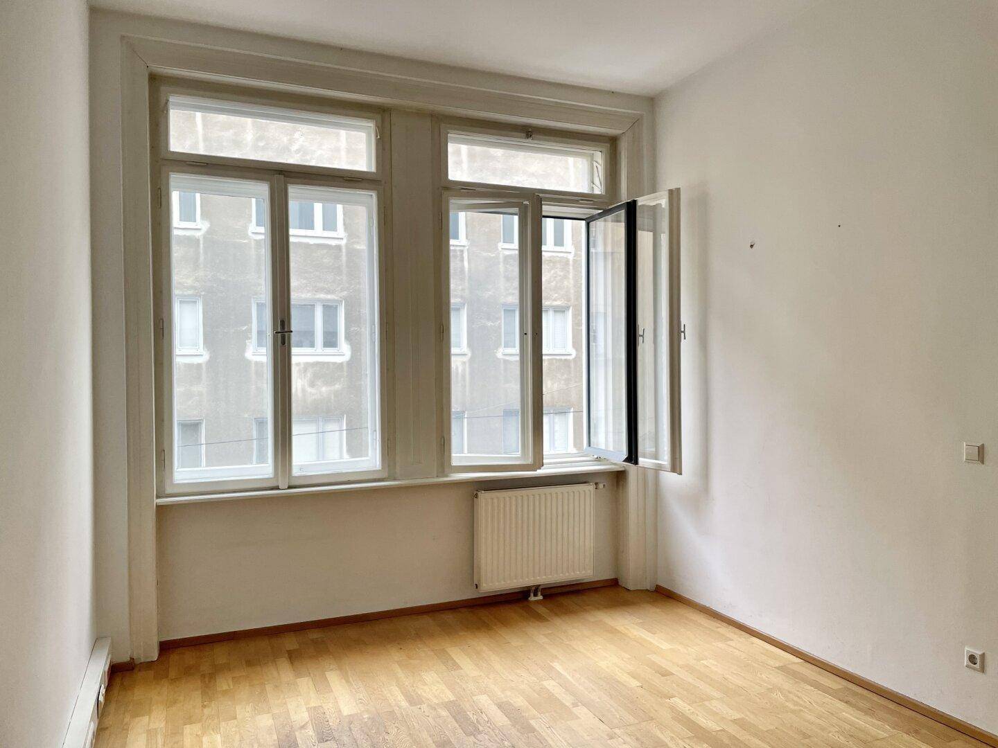 Leerer Raum mit geöffnetem Fenster, Parkettboden und Blick auf die gegenüberliegenden Gebäude.