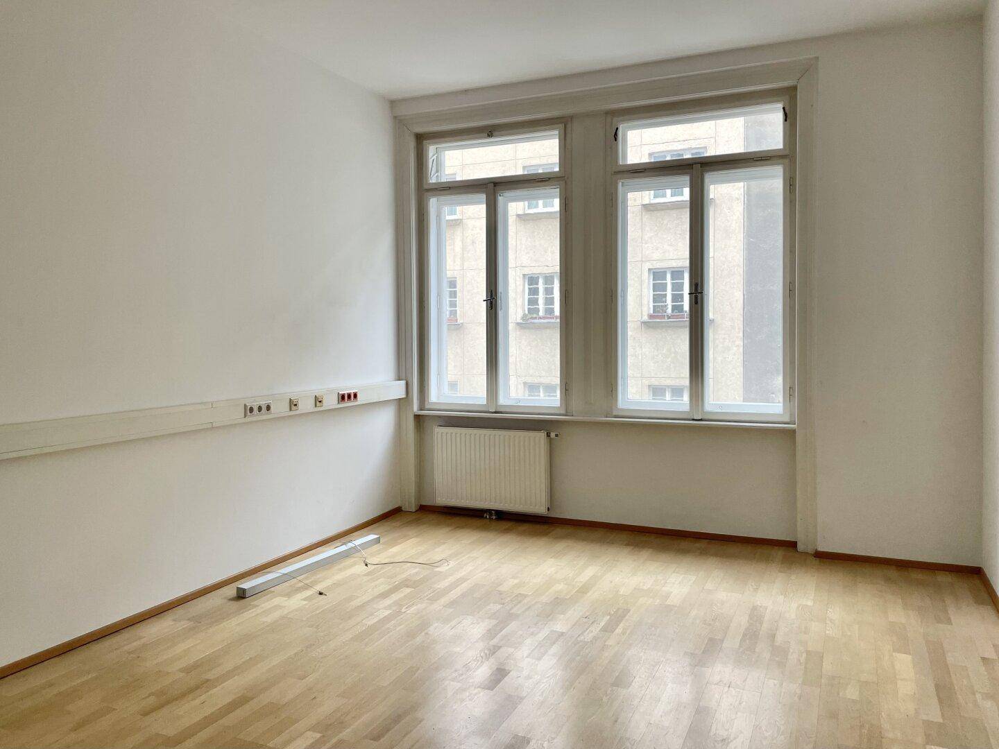 Heller Raum mit Parkettboden, großen Fenstern und einer Wand mit Steckdosenleiste.