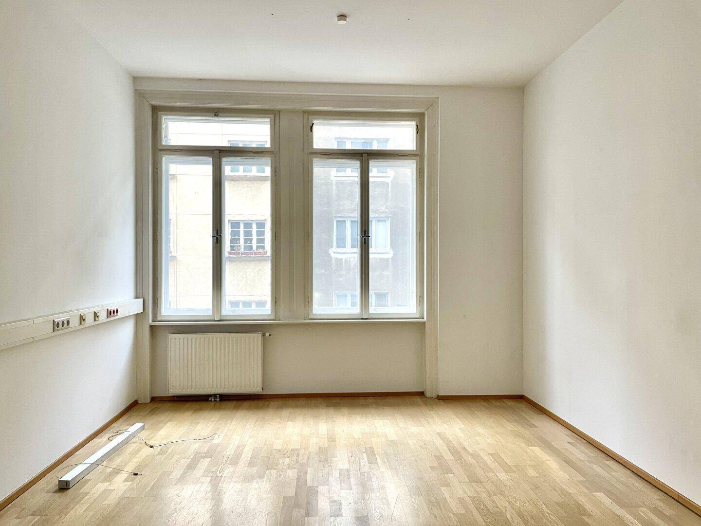 Leerer Raum mit Parkettboden, großen Fenstern und einer langen Steckdosenleiste an der Wand.