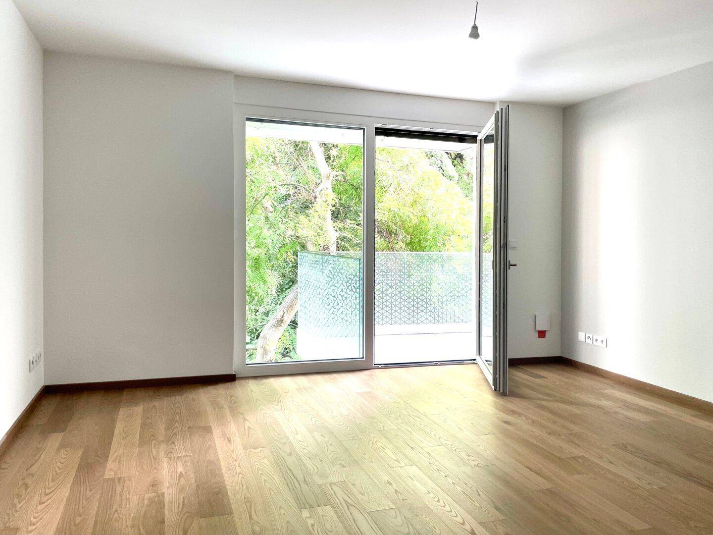 Heller, leerer Raum mit Holzboden und großen Fenstern, die Zugang zu einem Balkon bieten.