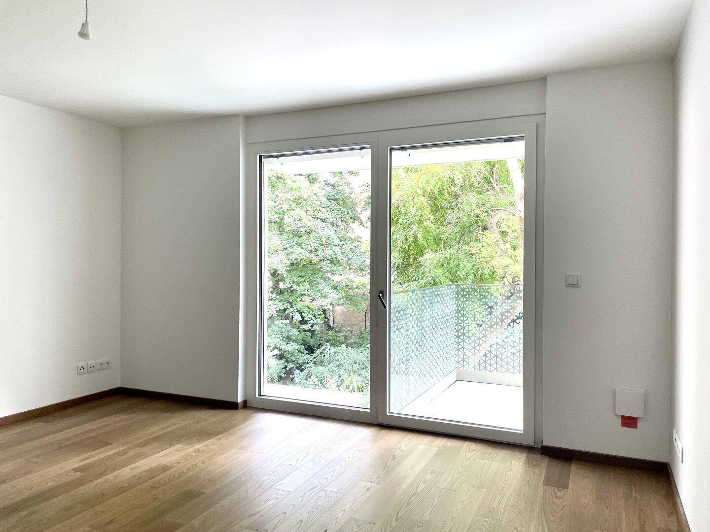 Geräumiger Raum mit hellem Holzboden und großem Fenster, das den Blick auf den Balkon freigibt.