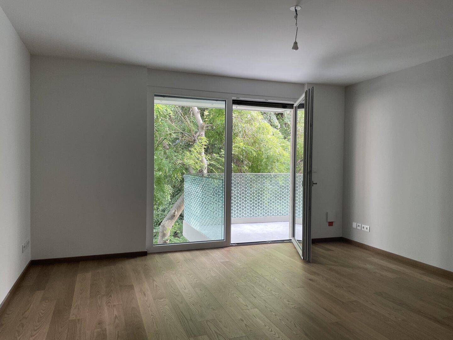 Heller, leerer Raum mit Holzboden und großen Fenstern, die Zugang zu einem Balkon bieten.