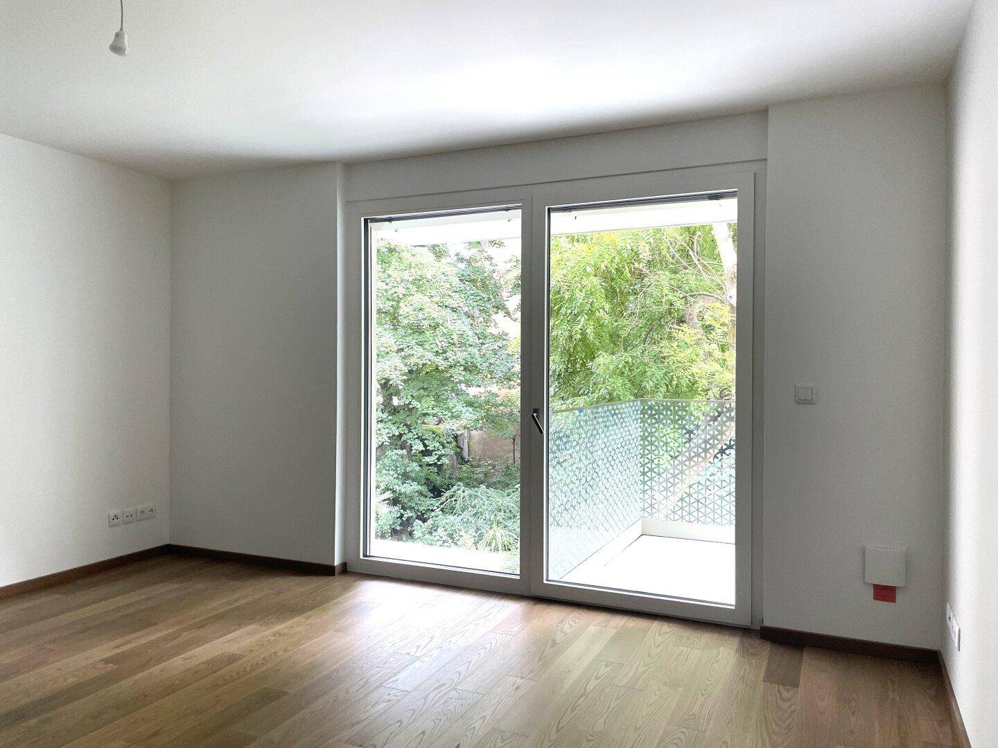 Geräumiger Raum mit hellem Holzboden und großem Fenster, das den Blick auf den Balkon freigibt.