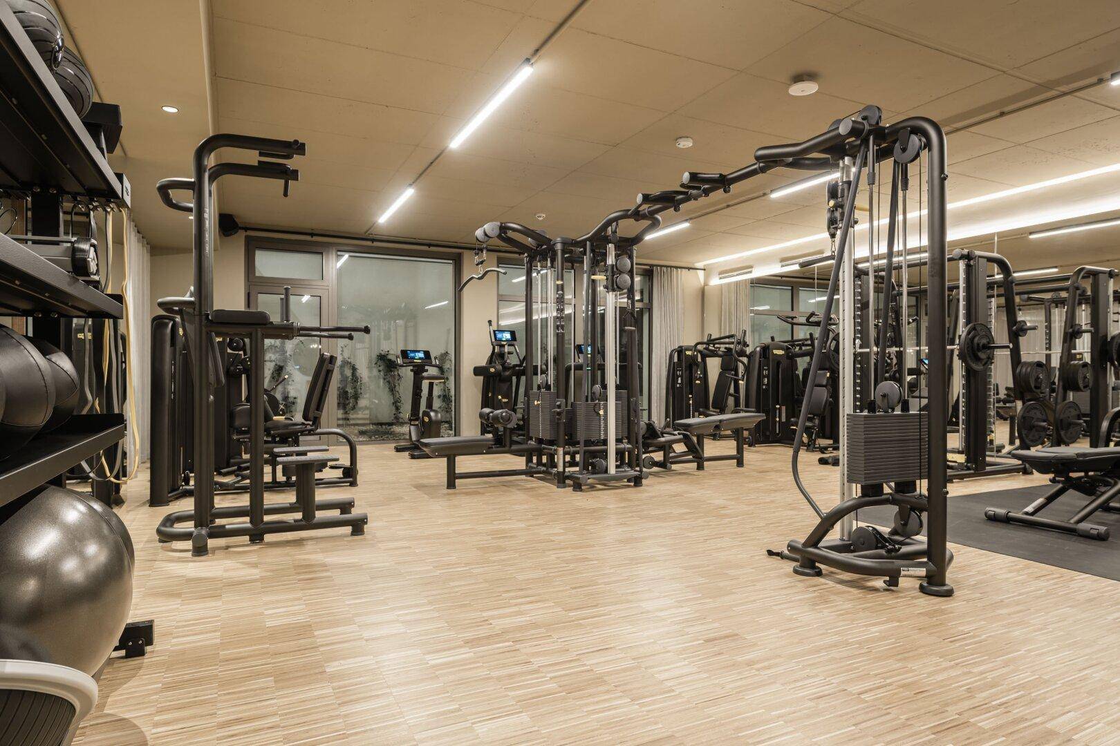 Moderner Fitnessraum mit vielfältigen Trainingsgeräten, großen Fenstern und hellem Laminatboden.