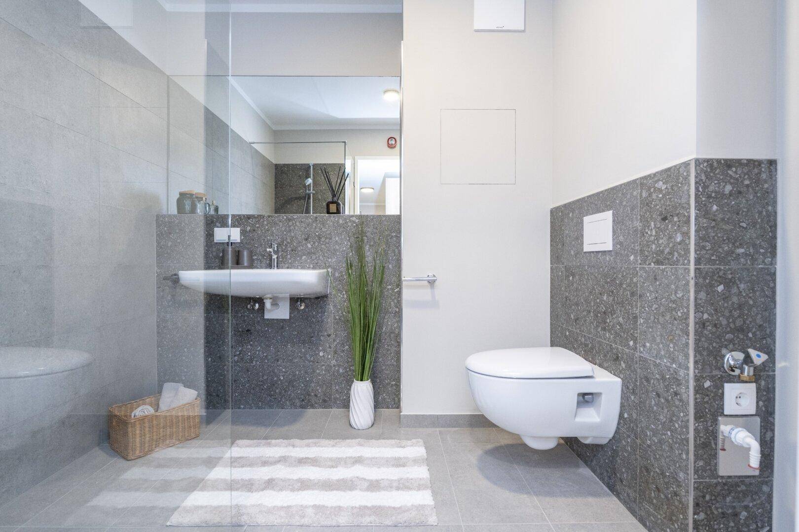 Modernes Badezimmer mit grauen Fliesen, wandhängendem WC und begehbarer Dusche.